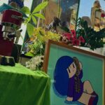 Red DeLonghi espresso machine surrounded by green plants and flowers, with a plush lion on top and a cartoon poster of a girl in purple on the side.