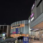 Night exterior of an AEON shopping center at night with a curved glass facade and lit pink AEON sign, visible taxi sign and bicycles nearby in front of the building.