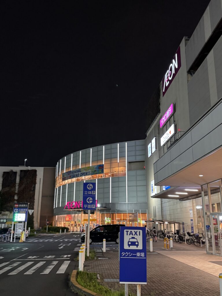 Night exterior of an AEON shopping center at night with a curved glass facade and lit pink AEON sign, visible taxi sign and bicycles nearby in front of the building.