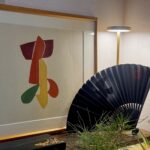 Framed abstract artwork with red, yellow, and green shapes, next to a black folding fan on a wooden surface with greenery nearby.