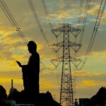 Silhouette of a Buddha statue in an urban street at sunset, with a large electricity pylon and power lines in the background.